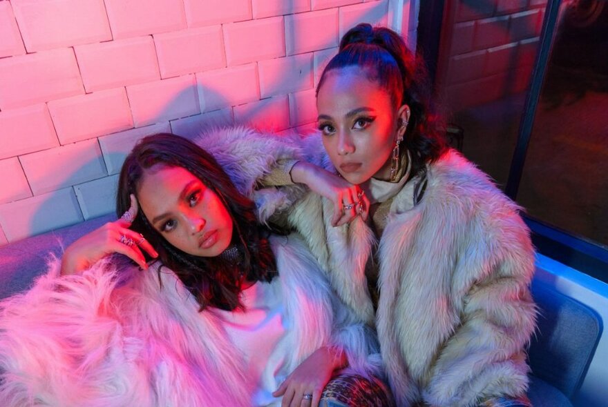 Two women looking sullen and bored in furry jackets on a sofa in a room with a pinkish glow. 