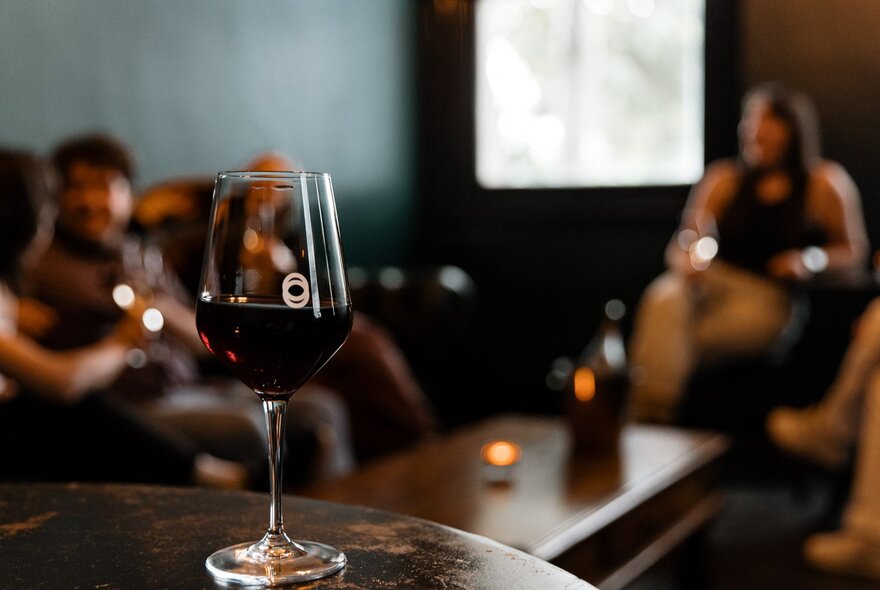 A glass of red wine on a round wooden table in sharp focus, people seated around a coffee table blurred in the background.