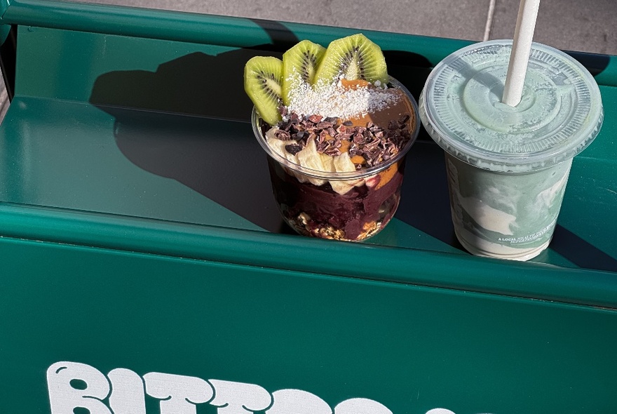 A smoothie in a takeaway clear plastic cup and an acai bowl filled with sliced banana and kiwi fruit and chocolate bits.