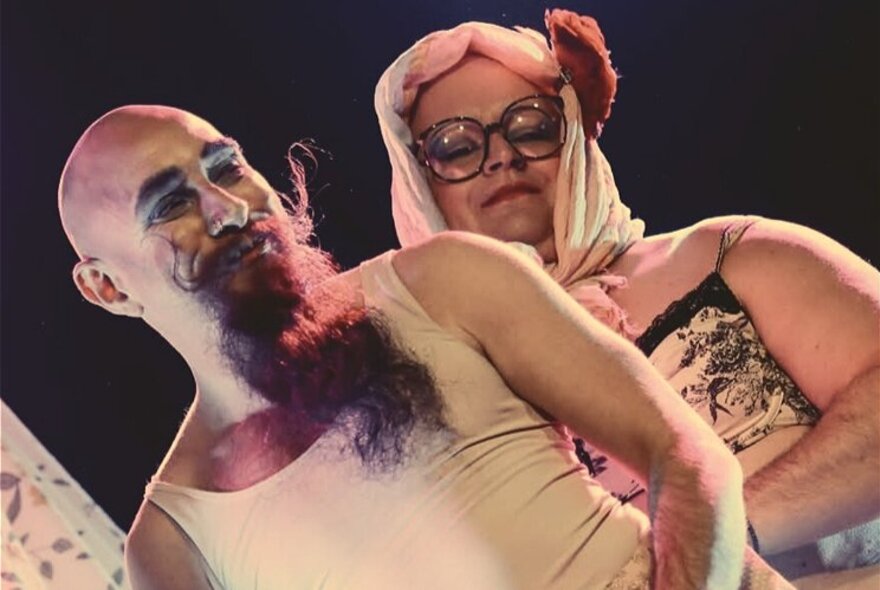 A man in a white singlet with a long beard and curly moustache with a woman behind him in a camisole and headscarf.