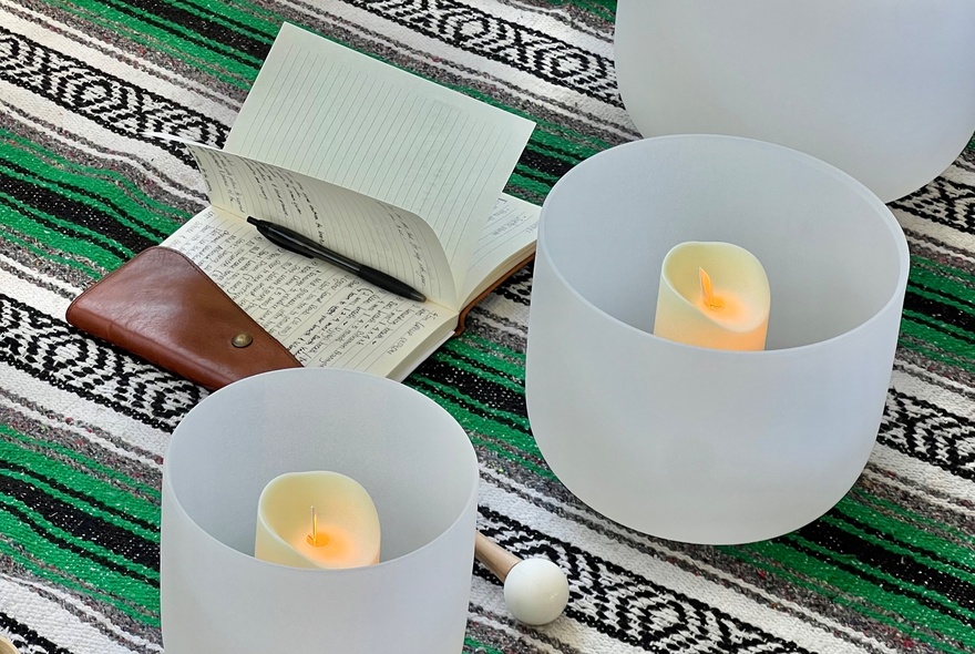Two white sound bowls with electric candles inside them, placed on a green and black striped cloth next to an open leatherbound notebook and pen.