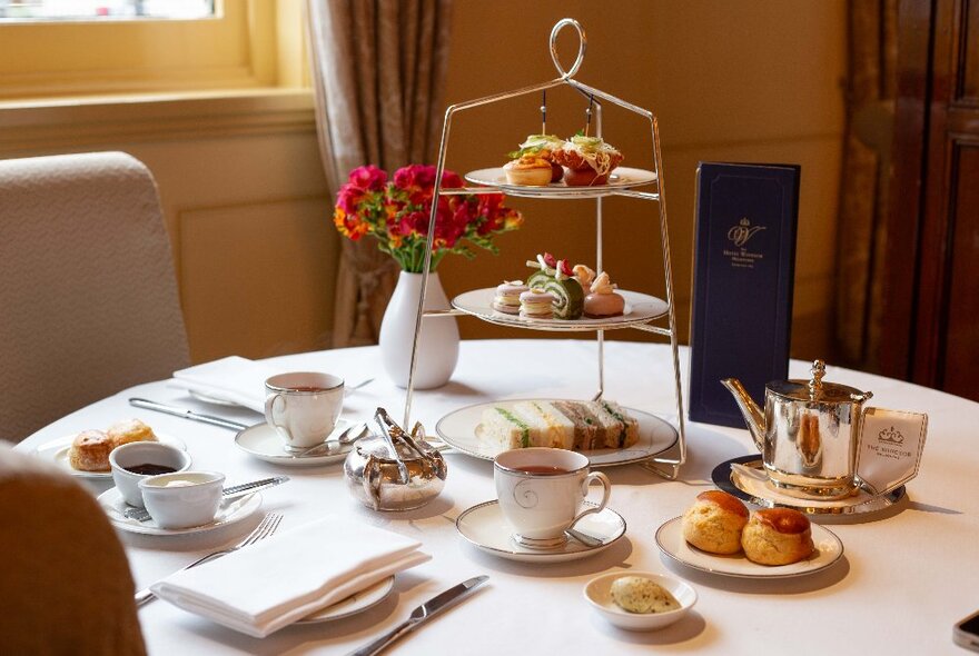 Afternoon tea with little plates of savoury and sweet food stacked up above each other on a three-tiered stand, on a table with pots of tea, cups and flowers.