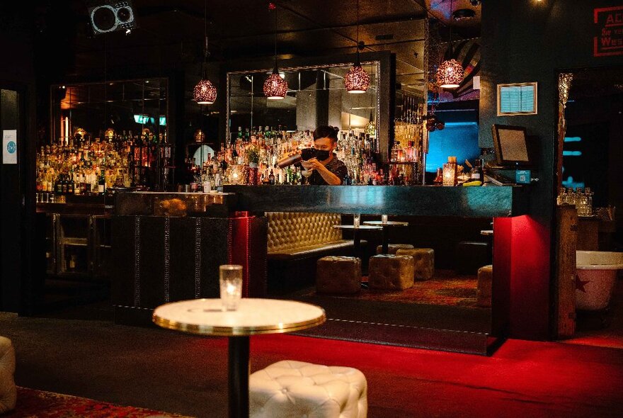 Intimate cocktail bar interior with tables and chairs, red carpet and bar tender behind a bottle-filled bar.