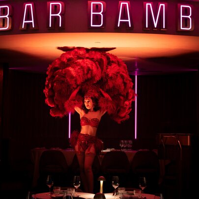 A burlesque dancer with huge feather fans performing under bar signage and in front of lamplit tables.
