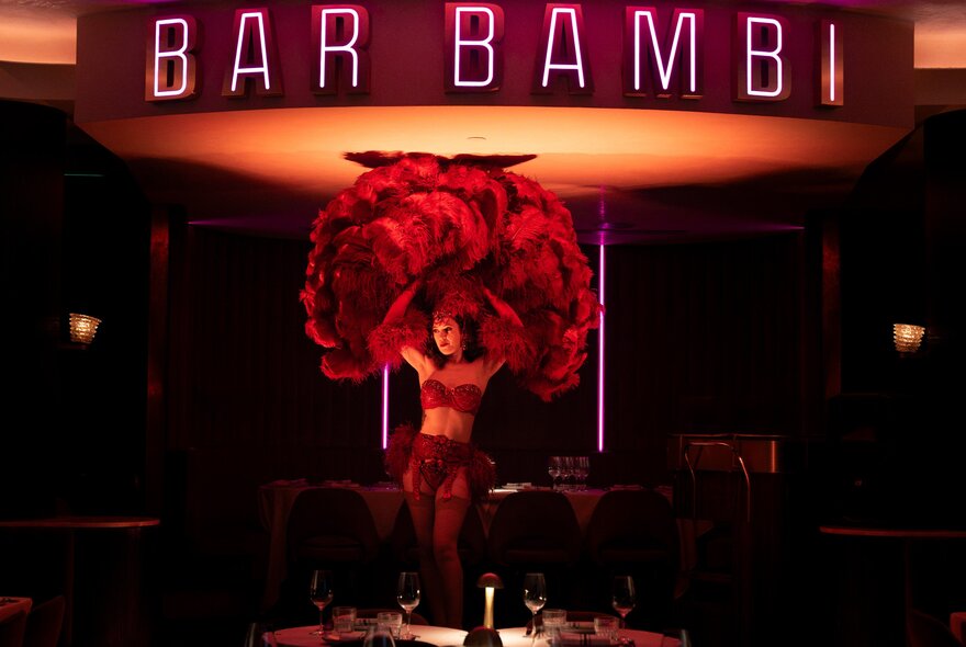 A burlesque dancer with huge feather fans performing under bar signage and in front of lamplit tables.