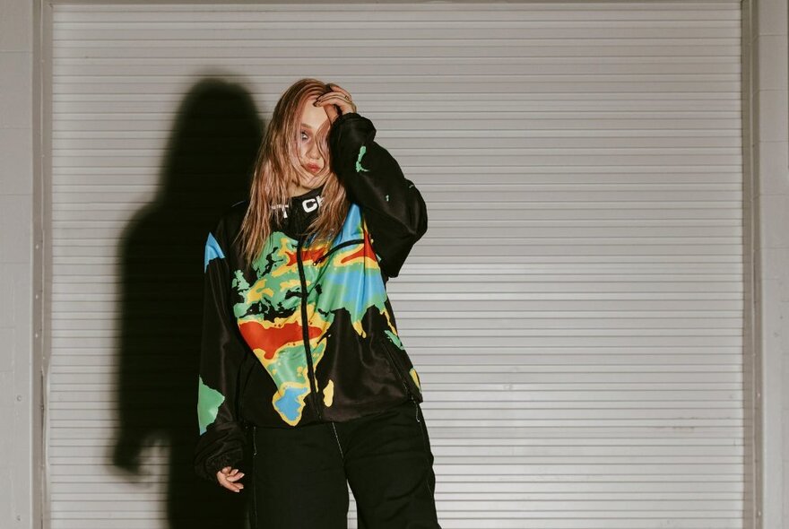 Singer George Alice, wearing a multi coloured top and black pants, standing in front of a roller shutter.