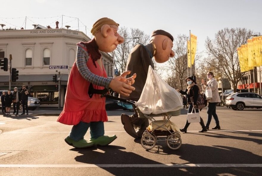 Two oversize giant puppets performing as roaming entertainers, walking along a city street, one of them pushing a pram.