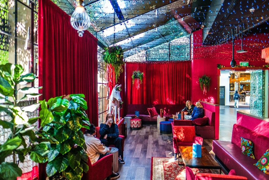 A colourful interior of a Mexican restaurant with hanging plants and fairy lights, and velvet red curtains, couches and pillows.