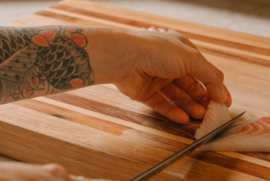 A hand and forearm with an intricate koi fish tattoo, slicing raw white fish with a sharp knife.