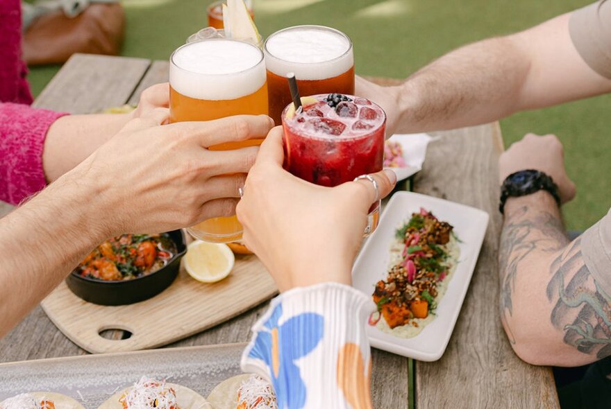 Four hands bringing four drinks in glasses together in a clinking motion over a table of food.