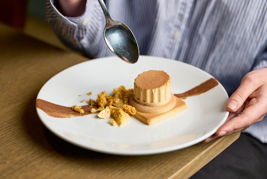 A spoon poised over a dessert plate with a pudding resembling the base of a column and crunchy rubble.