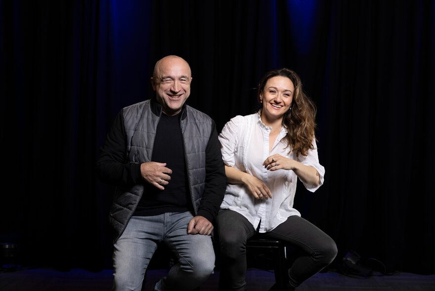 Two smiling actors in casual clothes seated on stools on a theatre stage.