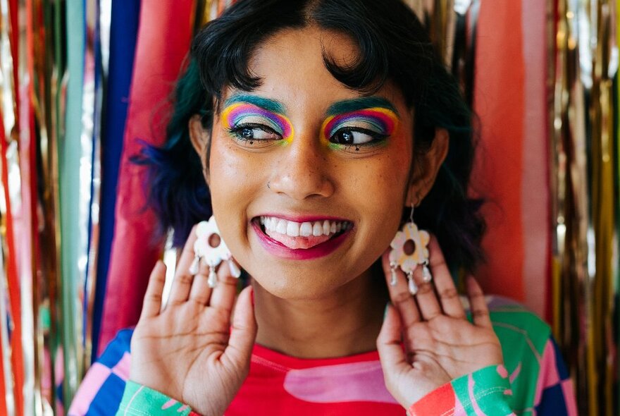 A young woman wearing a colourful top and rainbow eye makeup with her hands behind her white flower earrings, in front of a colourful stripy background.