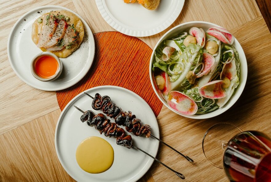 Looking down at a restaurant table with several dishes of food  and a glass of wine.