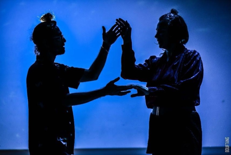 Two performers silhouetted on a stage, their hands touching, against a blue background.