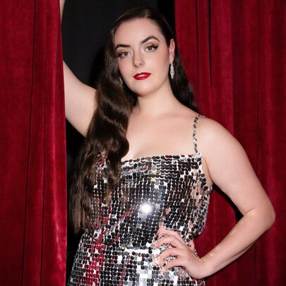 A burlesque dancer in a mirrored costume with long hair over one shoulder and a hand on her hip holding a red curtain aside. 