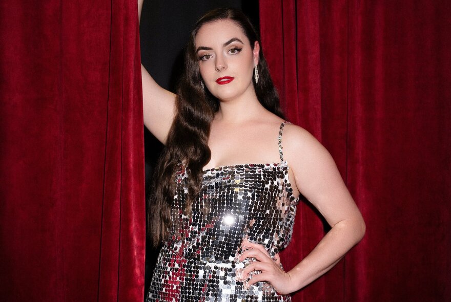 A burlesque dancer in a mirrored costume with long hair over one shoulder and a hand on her hip holding a red curtain aside. 