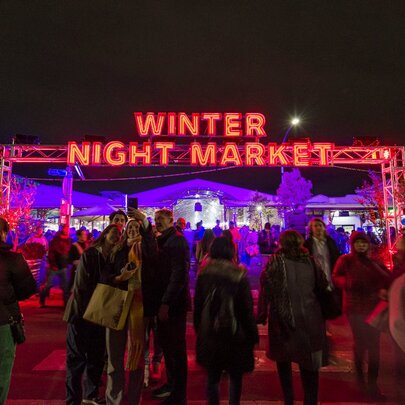 People walking towards a red illuminated archway that reads Winter Night Market. 
