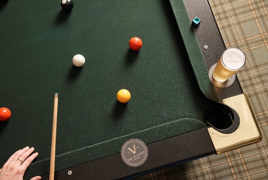 A dark green pool table seen from above with colourful billiard balls on it, a glass of beer on a coaster on the outside of the pool table.