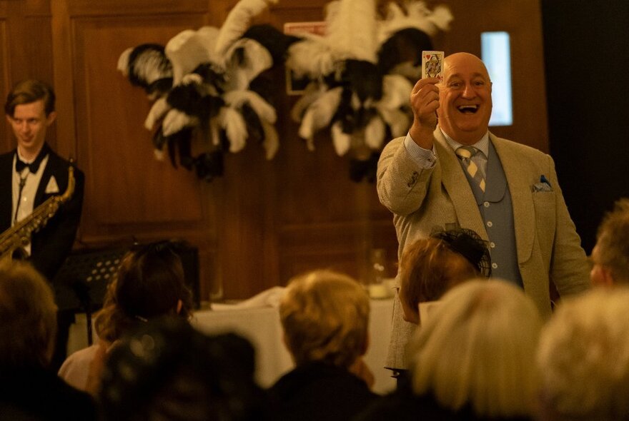 A man wearing a suit performing a card trick in front of a seated audience inside a wood panelled dining room.