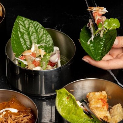 A person assembling a leaf wrap with various ingredients from metal bowls, including vegetables, meat, and a sauce, using chopsticks on a black table.