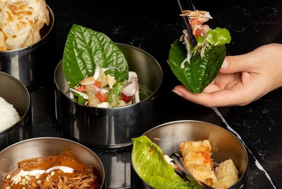 A person assembling a leaf wrap with various ingredients from metal bowls, including vegetables, meat, and a sauce, using chopsticks on a black table.