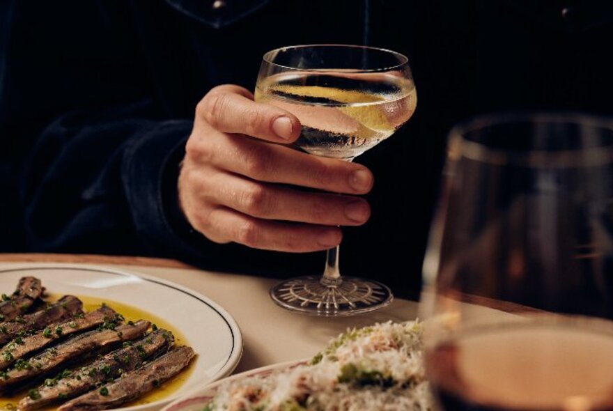 A hand holding a glass of white wine with various plates around the table. 