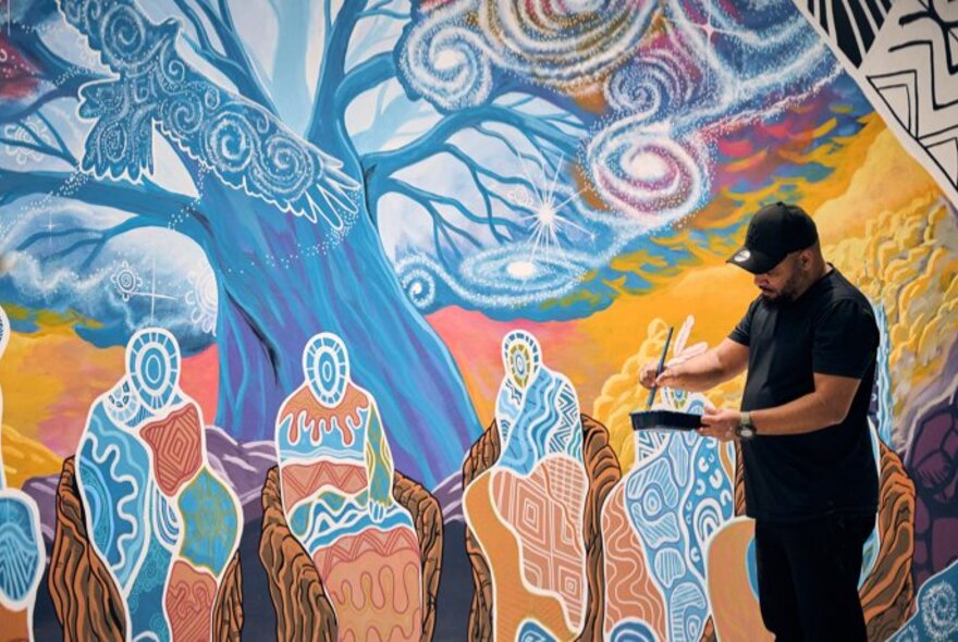 A gallery space featuring an indigenous mural artwork on the wall, an Indigenous man holding an easel and a paint brush in front of it.