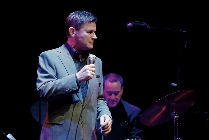 Perfomer Gregg Arthur holding a microphone performing on stage with musicians in the background.