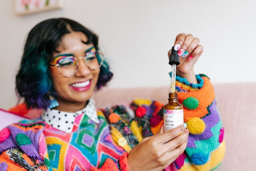 A young woman wearing a vibrant, colourful jumper with bobbles and rainbow eye makeup lifting a dropper from a small bottle.