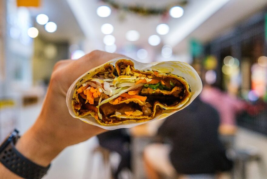 A hand holding up a filled Chinese crepe with an out of focus food court in the background.