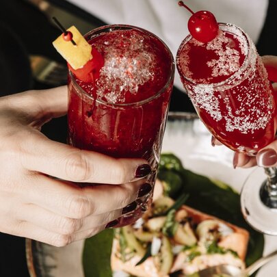 Two people toast with tall red cocktails over a table with a plate of food.