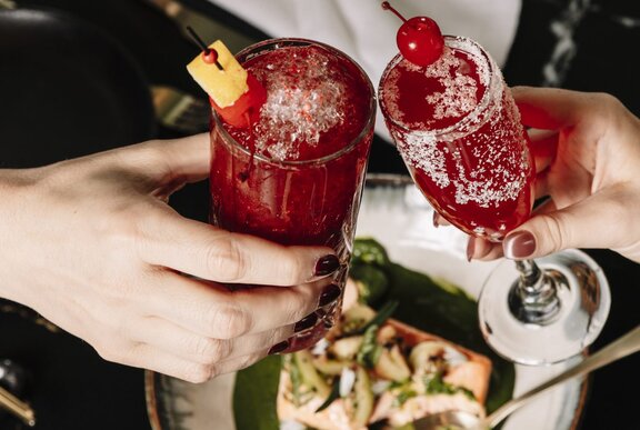 Two people toast with tall red cocktails over a table with a plate of food.