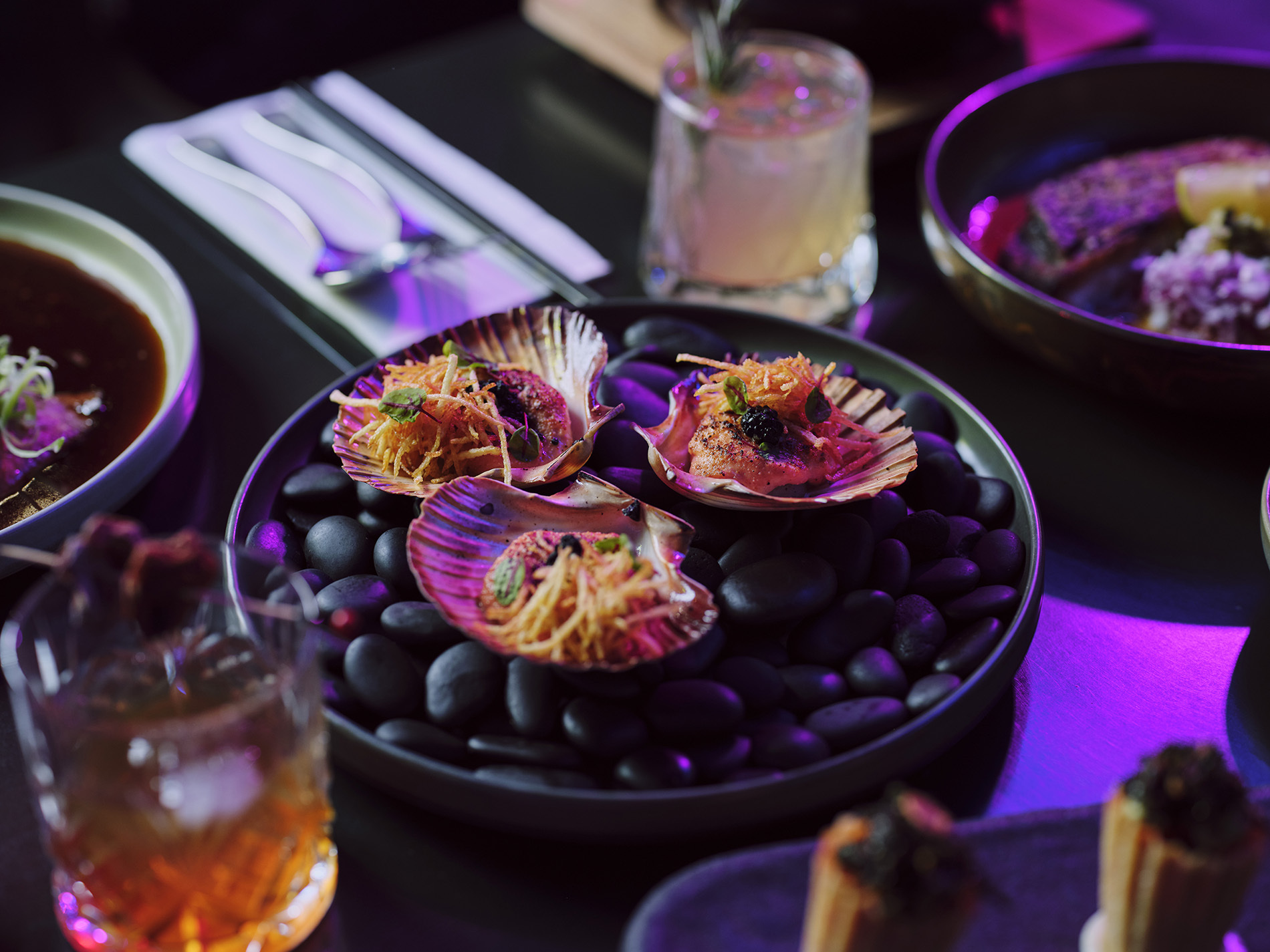 A plate of scallops surrounded by cocktails and other dishes.