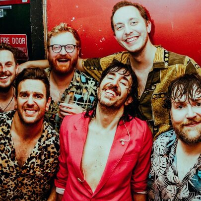 Six members of a rock and roll band all wearing colourful shirts and smiling.