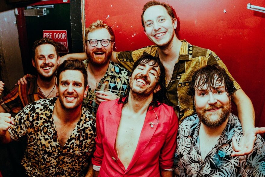 Six members of a rock and roll band all wearing colourful shirts and smiling.