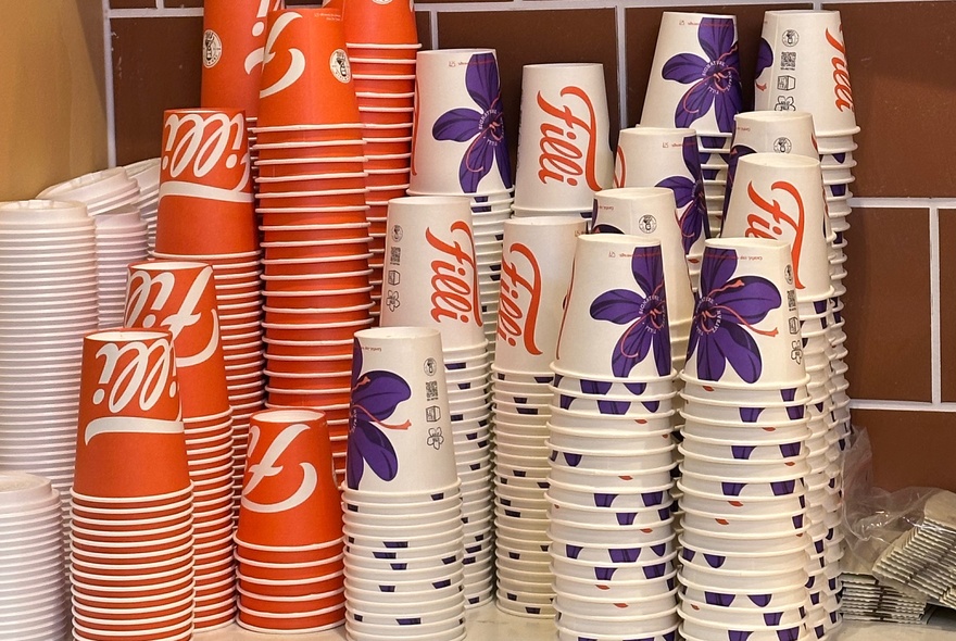 Two stacks of orange disposable takeaway cups and four stacks of white takeaway cups with purple flowers balanced on a bench with stacks of plastic lids next to them.