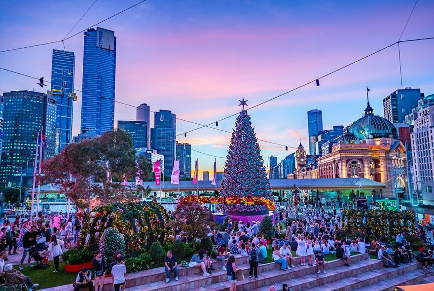 Crowds gathered around the Christmas tree at Fed Square at dusk, with Flinders Street Station and city buildings in background.