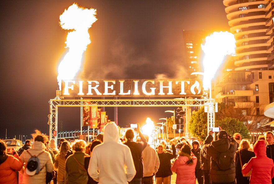 A crowd of people are walking under an archway of flames with a sign saying firelight. 