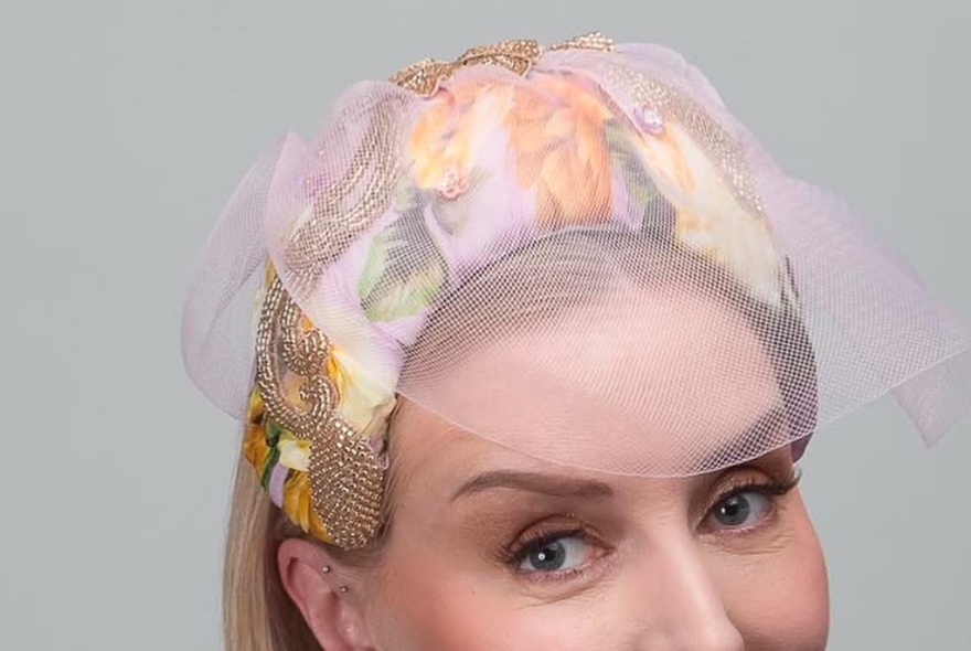 A headshot of a woman wearing a peachy patterned fascinator with mesh over her forehead.