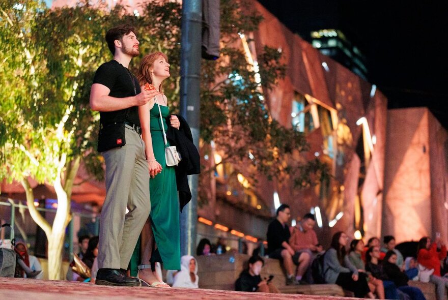 Two people standing and holding hands, watching a movie at Fed Square at night.