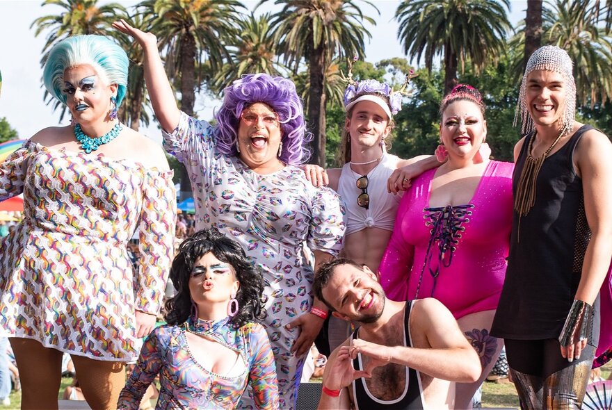 A group of people, some in drag with bright wigs and bold make-up, smiling while posing for a photo outdoors at Alexandra Gardens during Midsumma Carnival.