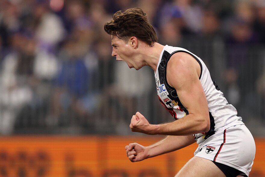 A St Kilda AFL player celebrating on the field with both his fists clenched, blurred fans in the background.