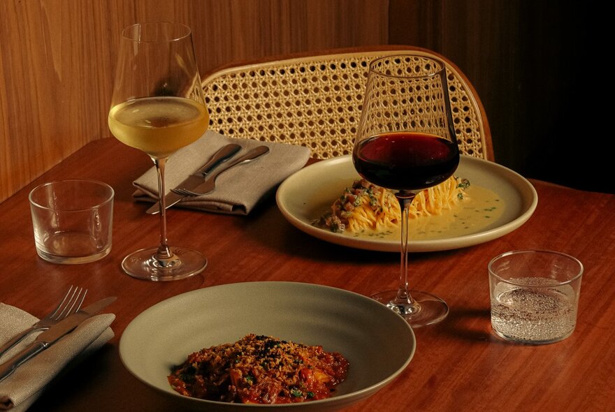 Two plates of pasta on a wooden dining table with two glasses of wine, cutlery and napkins.