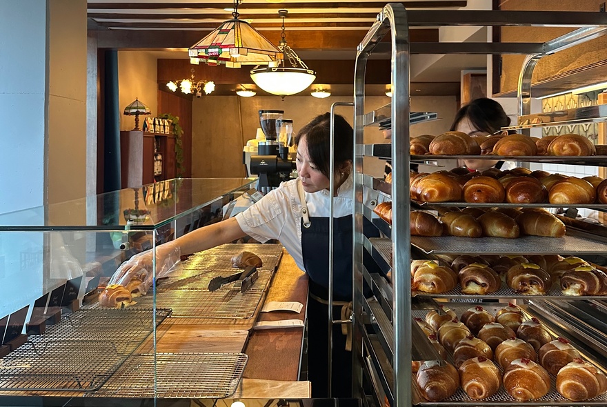 Baker moving freshly baked croissants from trays onto a glass counter in a cafe with pendant lights.