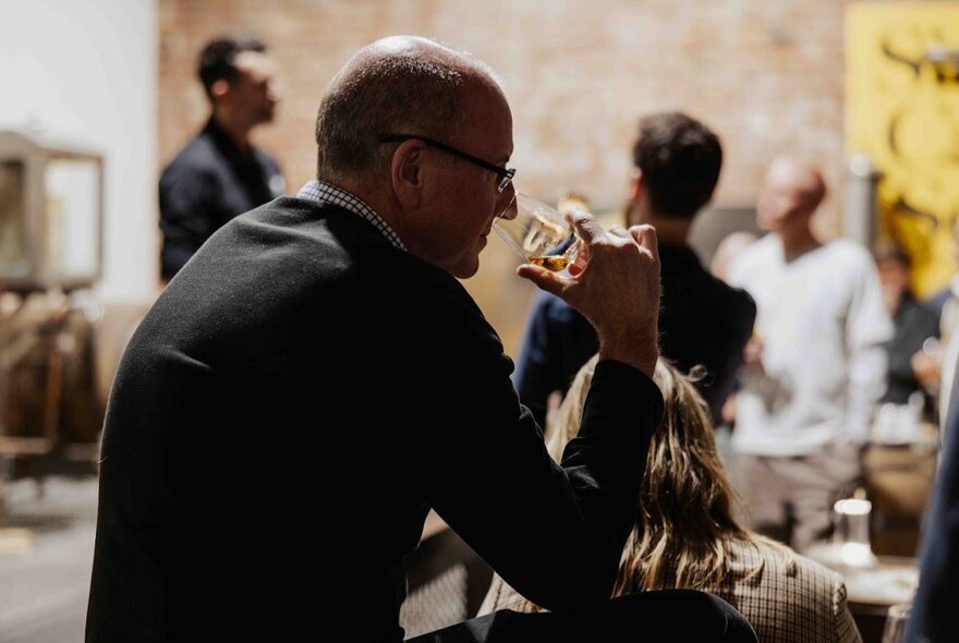 A man seated at a table tasting whisky in a large space, figures in the background.