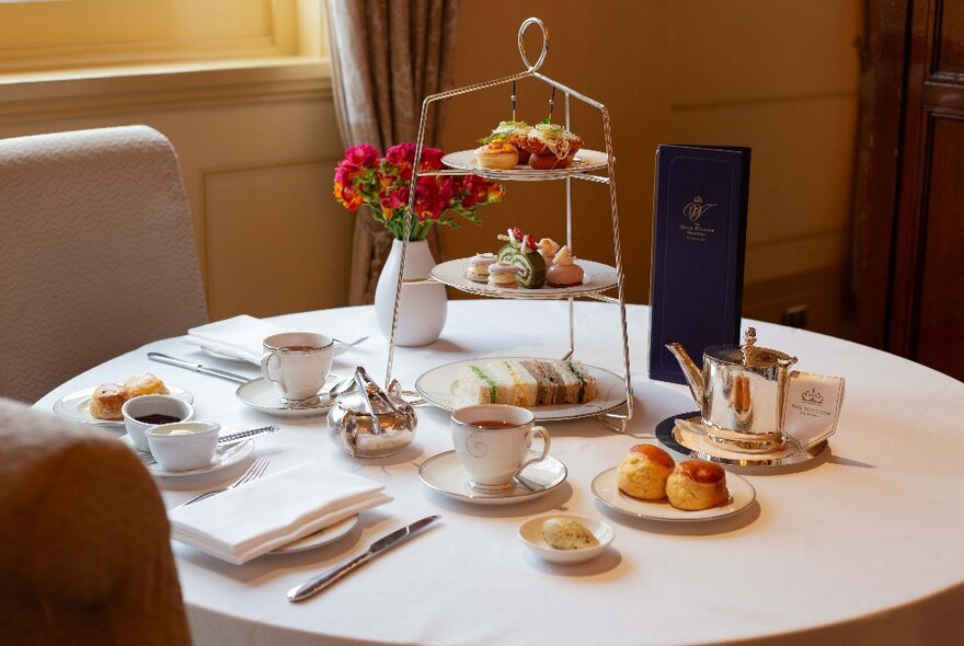 A tiered platter of afternoon tea sweet treats, with fine china tea cups and saucers, a stainless steel teapot and white napkins.