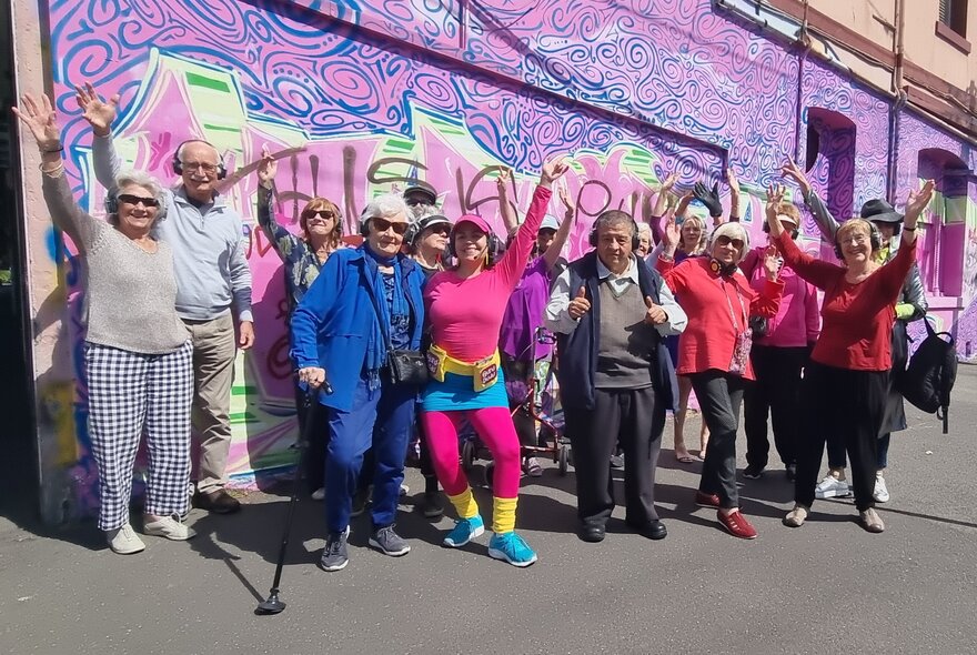 A group of older people all standing together in from of a graffitied wall, some with their hands in the air. 