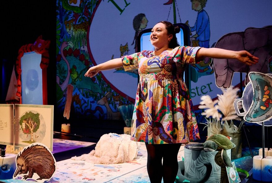 Performer standing with arms outstretched on stage next to animal props.