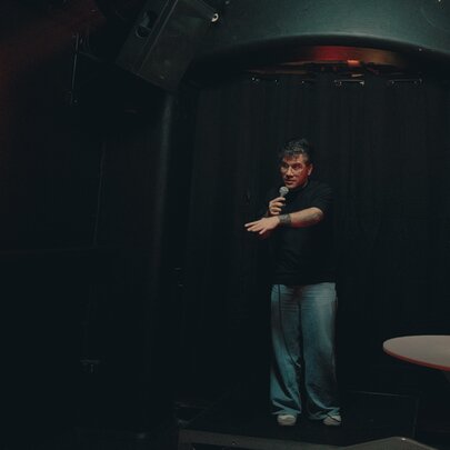 A dark image of a comedian on stage in a black t-shirt and jeans.